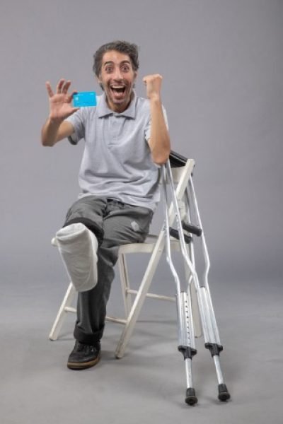 front-view-young-male-sitting-with-broken-foot-holding-blue-bank-card-grey-floor-pain-foot-broken-accident-leg-twist (Phone)